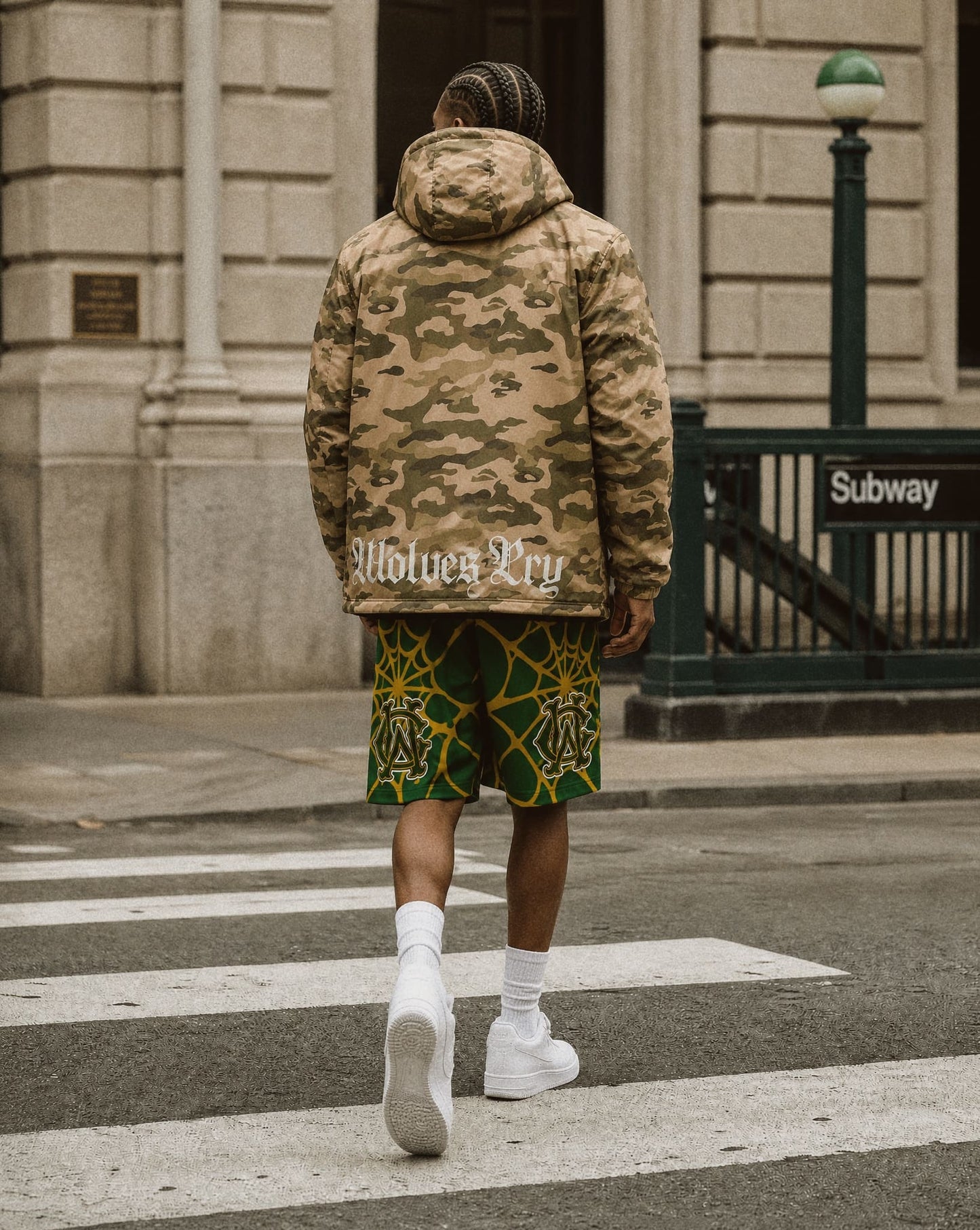 A man viewed from behind walking on a city crosswalk, wearing a beige and brown camouflage hooded jacket with 'wolves cry' text on the back, green shorts with a yellow geometric pattern, and white sneakers.