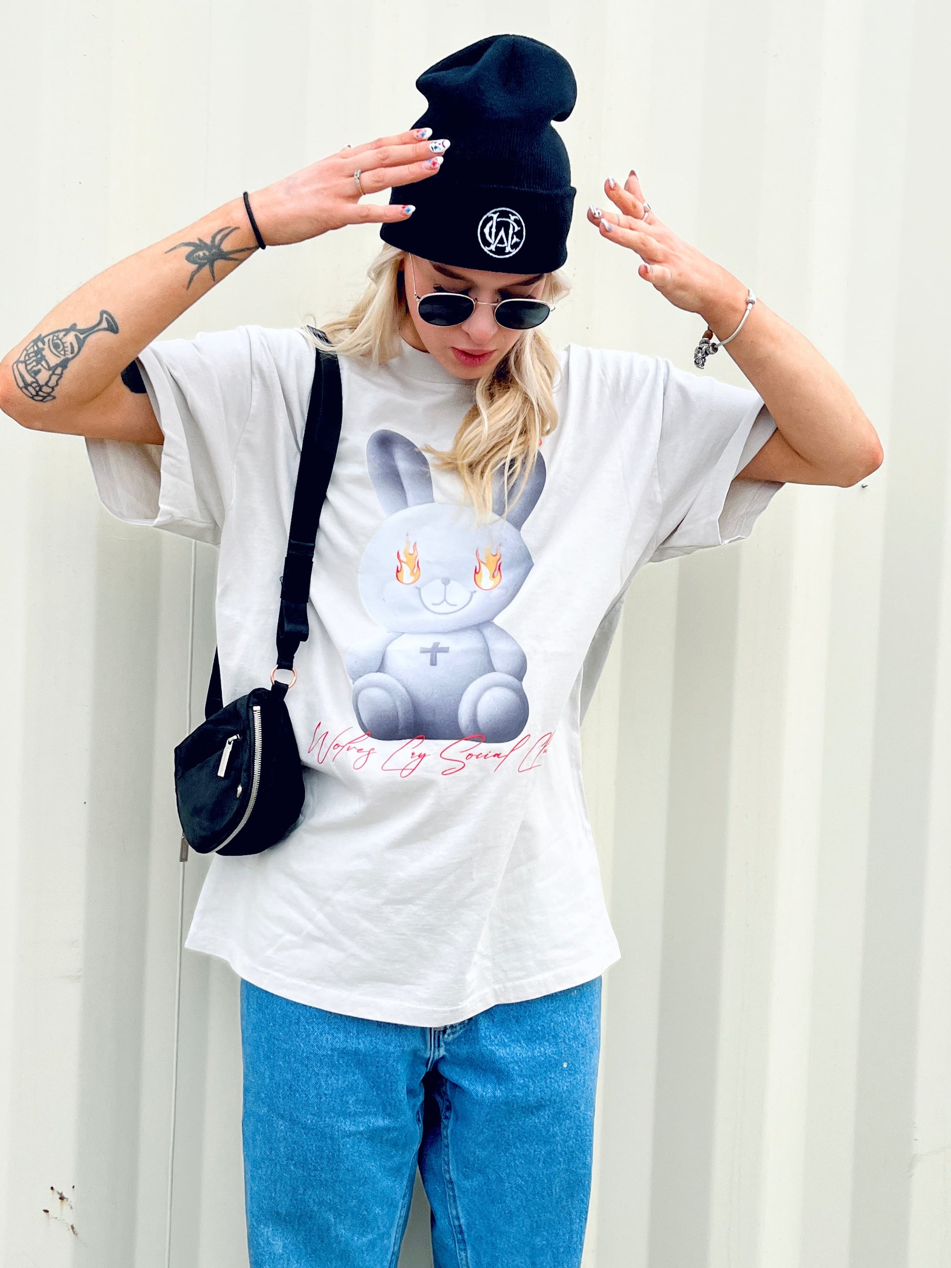 Model wearing the Wolves Cry Oversized Teddy Tee in bone off-white, featuring a graphic of a teddy engulfed in flames, styled with a black beanie and sunglasses.