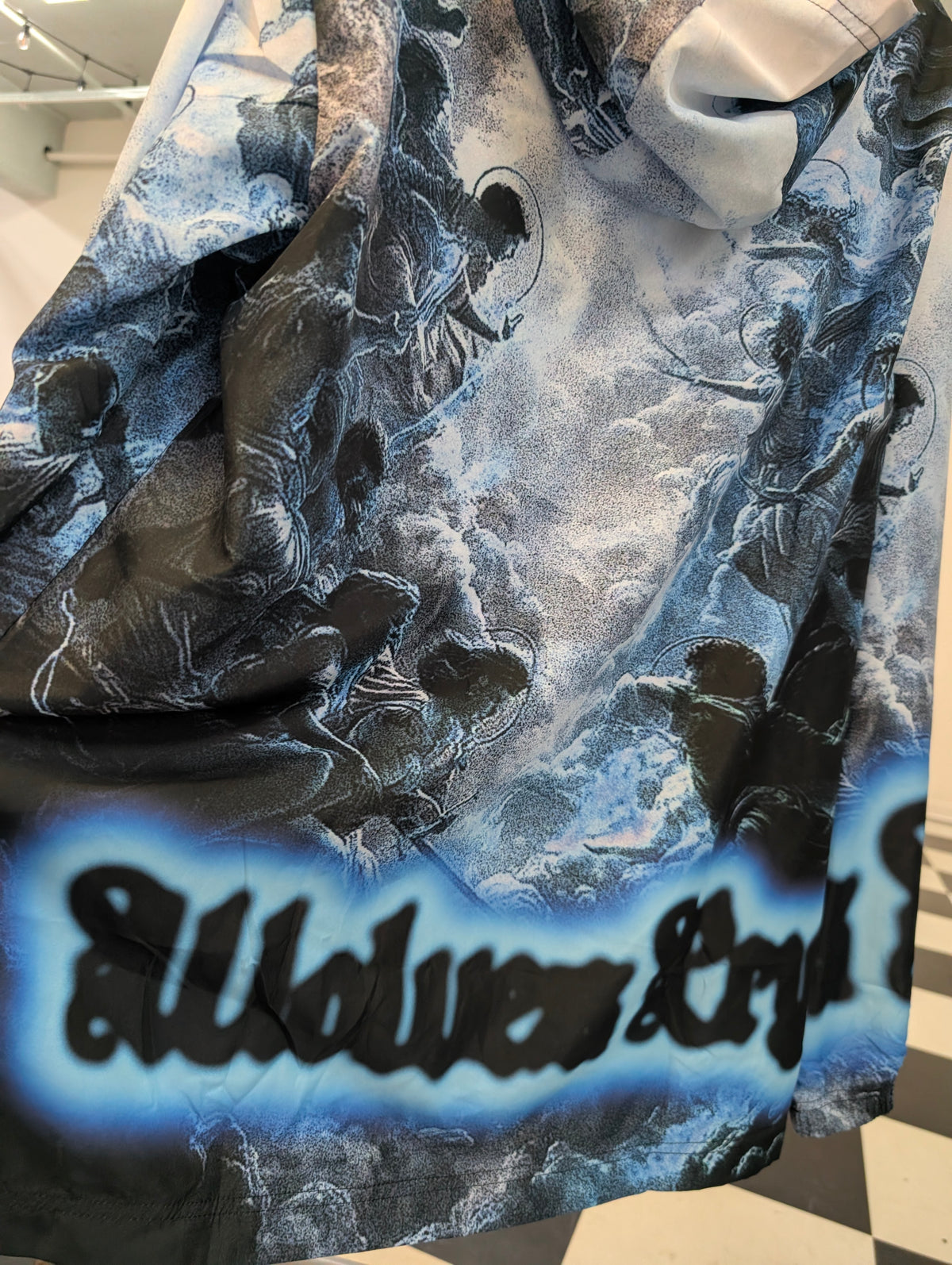 A close-up of a Wolves Cry Windbreaker graphic print with figures in black and white, set against a blue and gray cloudy sky. The bottom features a bold black gothic-style text graphic outlined in bright blue.