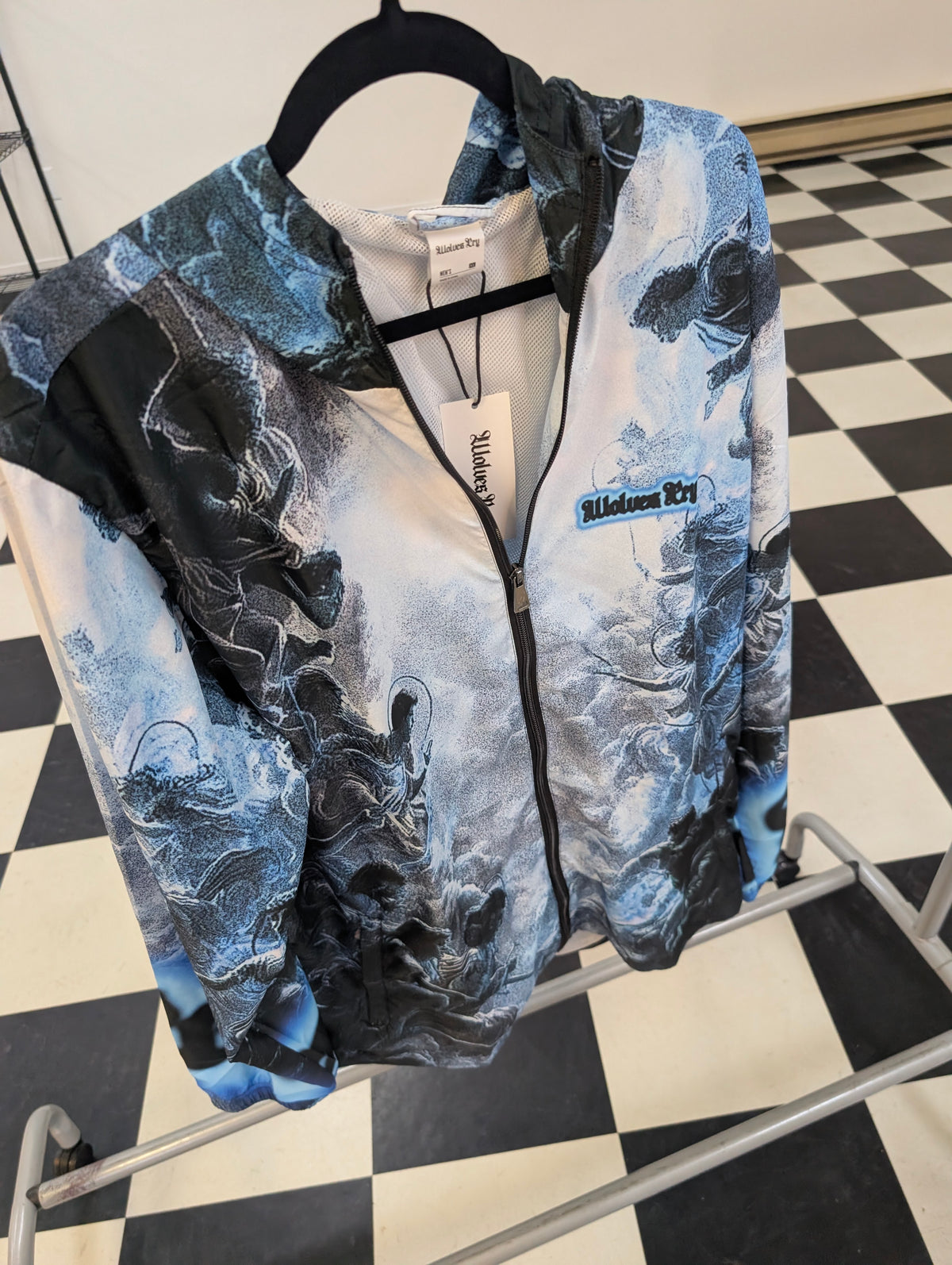 A Wolves Cry zip-up jacket with a blue and black abstract print, hanging on a black hanger on a clothing rack over a black and white checkered floor.