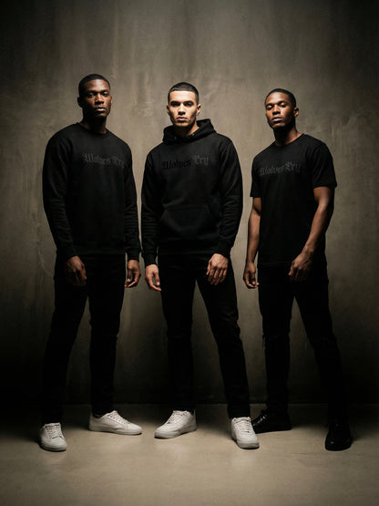 Three men wearing black hoodies and pants standing against a gray background
