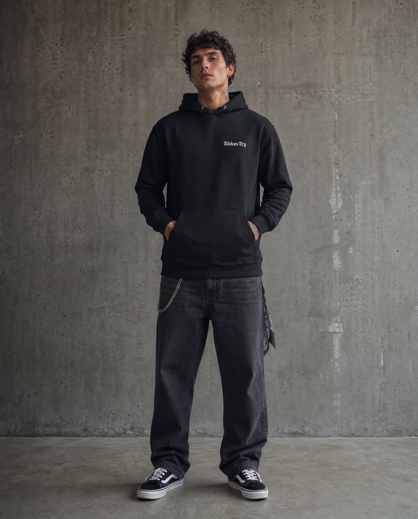 Man wearing a black hoodie with text and dark jeans standing against a gray concrete wall.