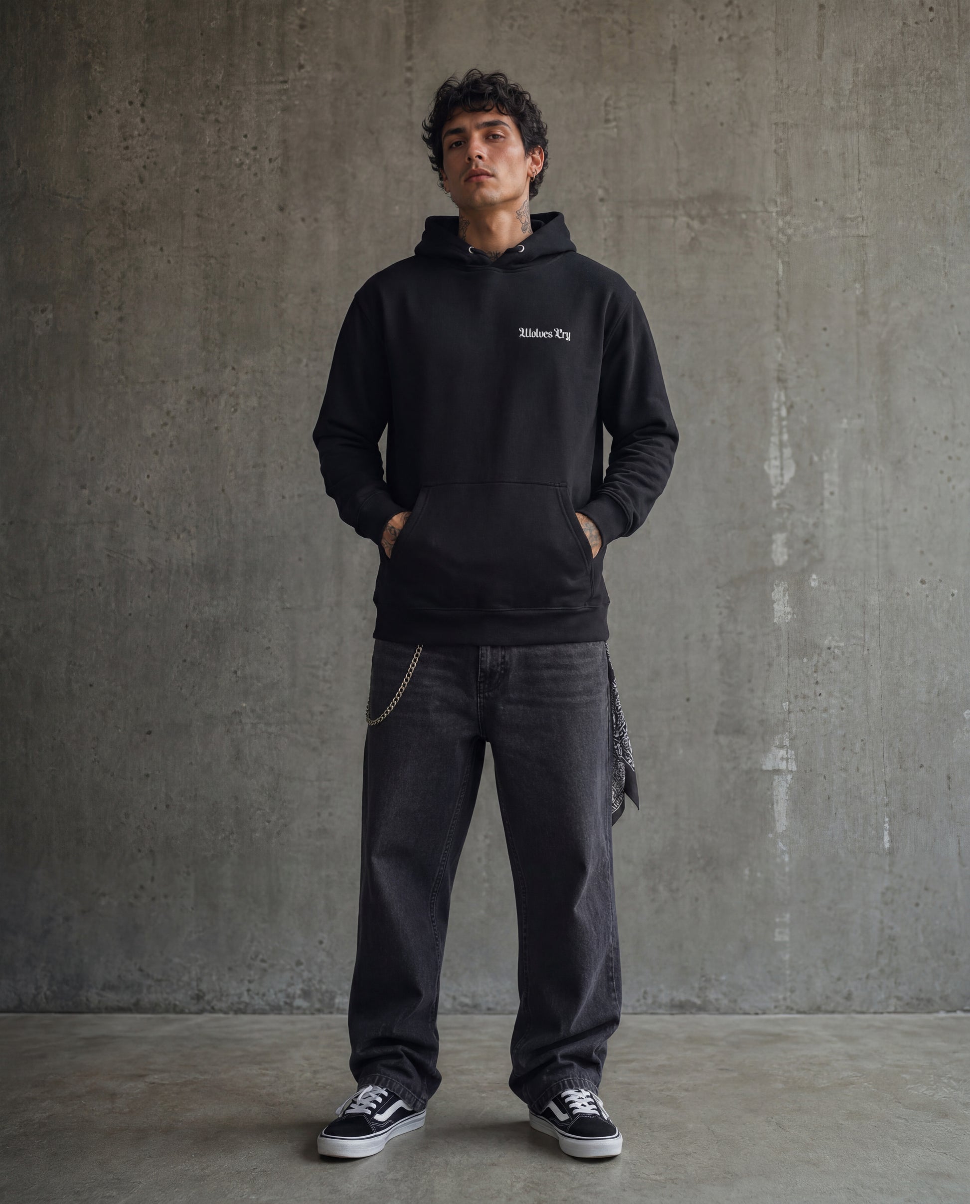 Man wearing a black hoodie with text and dark jeans standing against a gray concrete wall.