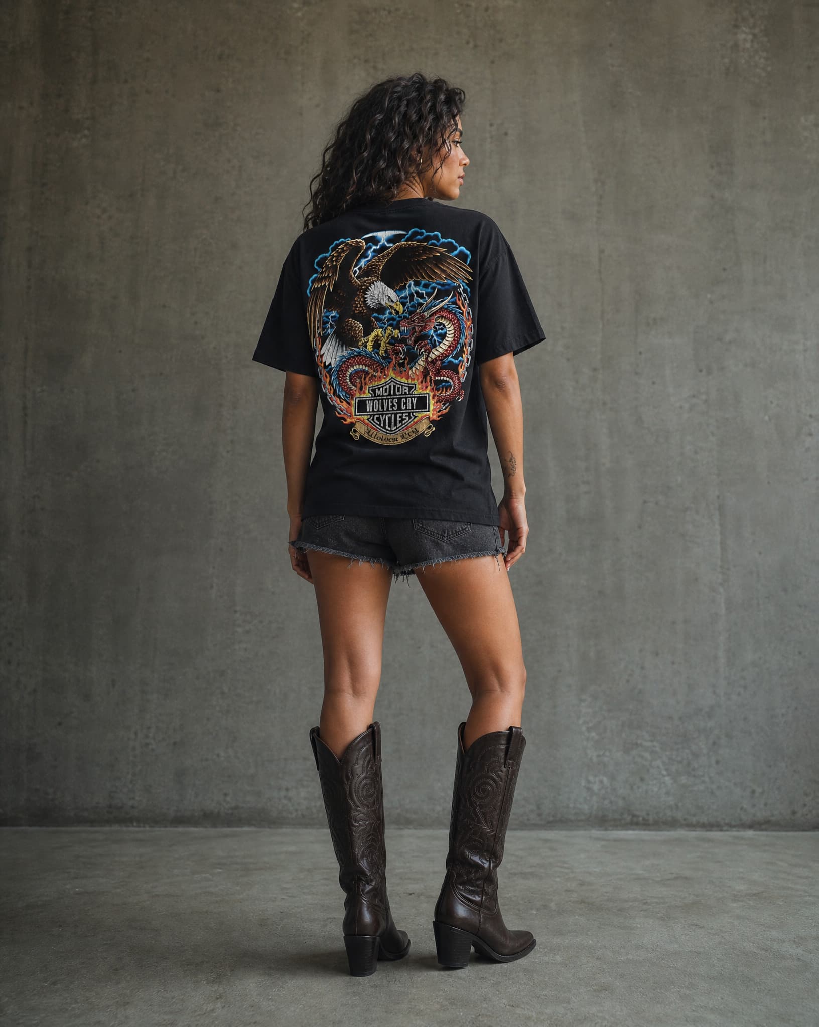 Person wearing a black t-shirt with a colorful graphic on the back, standing against a gray concrete wall.