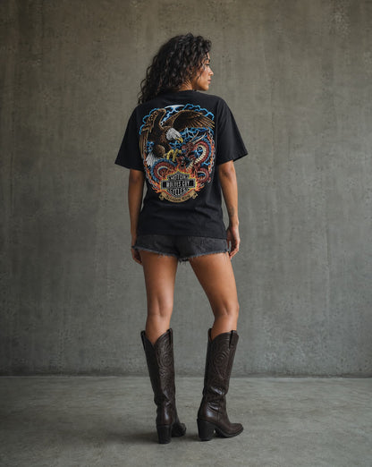 Person wearing a black t-shirt with a colorful graphic on the back, standing against a gray concrete wall.