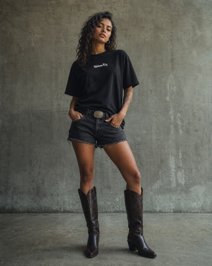 Woman wearing a black t-shirt, denim shorts, and brown knee-high boots against a gray concrete wall.