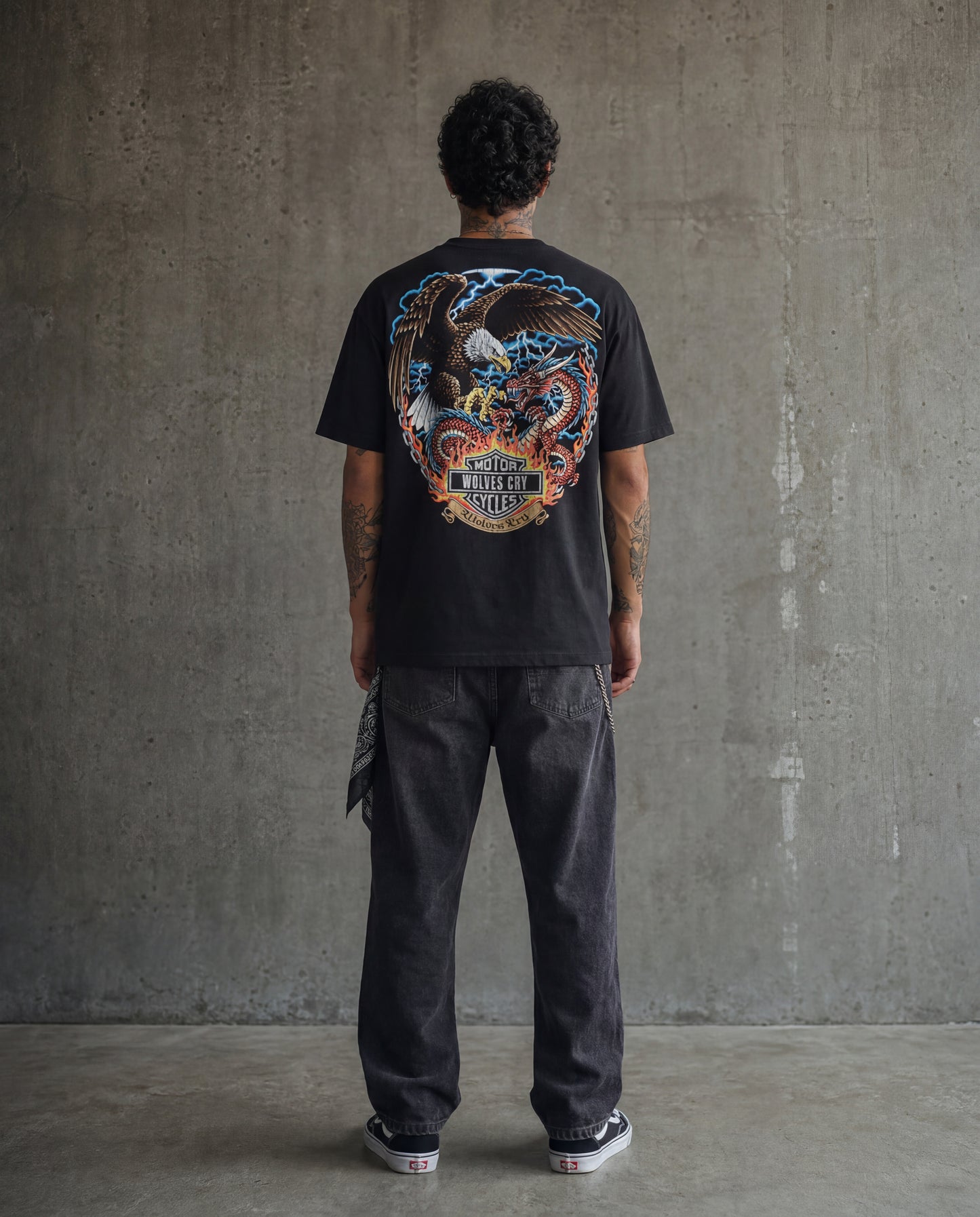 Person wearing a black t-shirt with a colorful graphic design on the back, standing against a gray concrete wall.