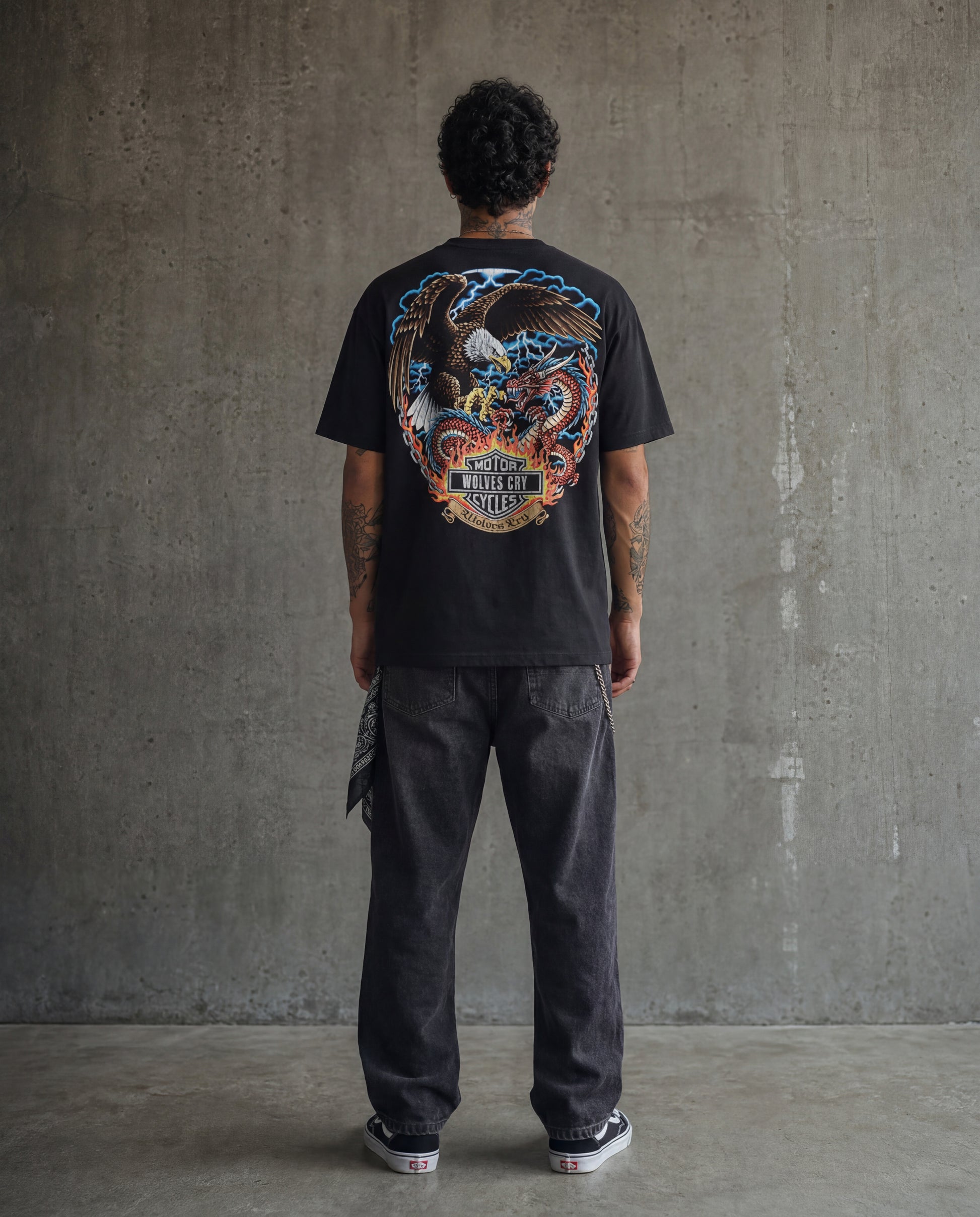 Person wearing a black t-shirt with a colorful graphic design on the back, standing against a gray concrete wall.