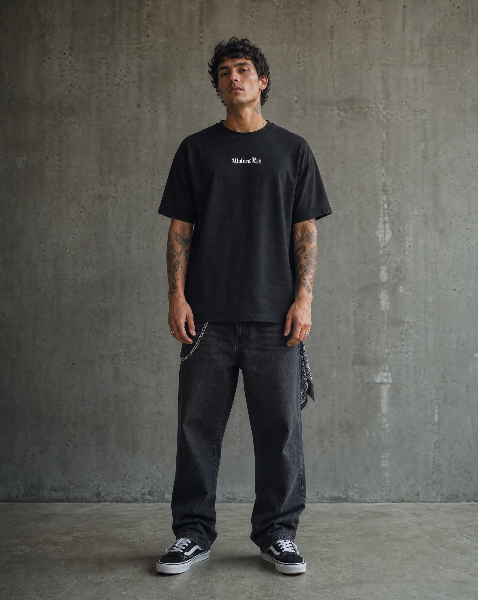 Man wearing a black t-shirt and dark jeans standing against a gray concrete wall.