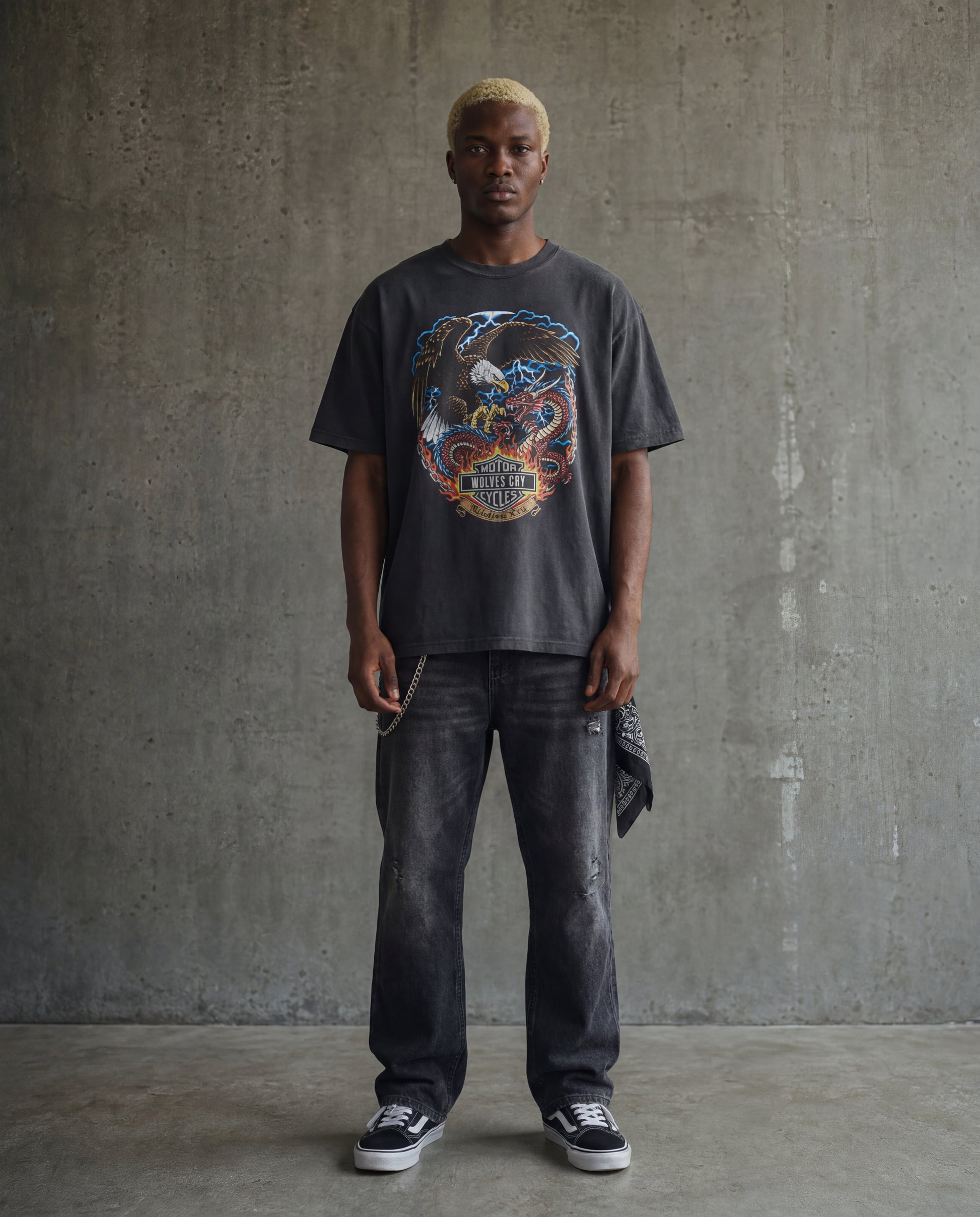 Man wearing a graphic t-shirt and dark jeans against a concrete wall.