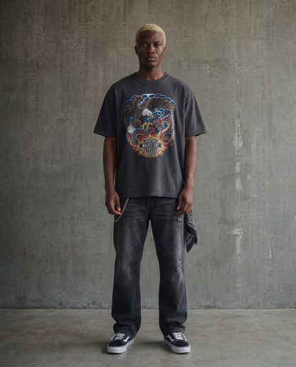 Man wearing a graphic t-shirt and dark jeans against a concrete wall.