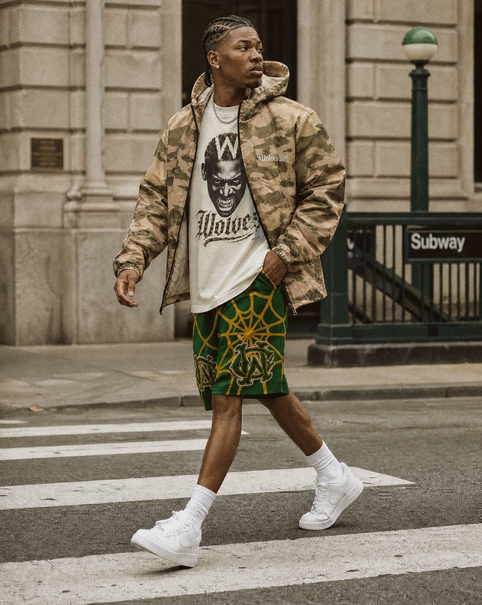A man walking on a city crosswalk wearing a camouflage ,wolves cry windbreaker jacket, a white graphic t-shirt, green and yellow patterned shorts, and white sneakers.