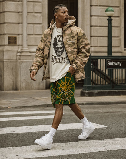 A man walking on a city crosswalk wearing a camouflage ,wolves cry windbreaker jacket, a white graphic t-shirt, green and yellow patterned shorts, and white sneakers.