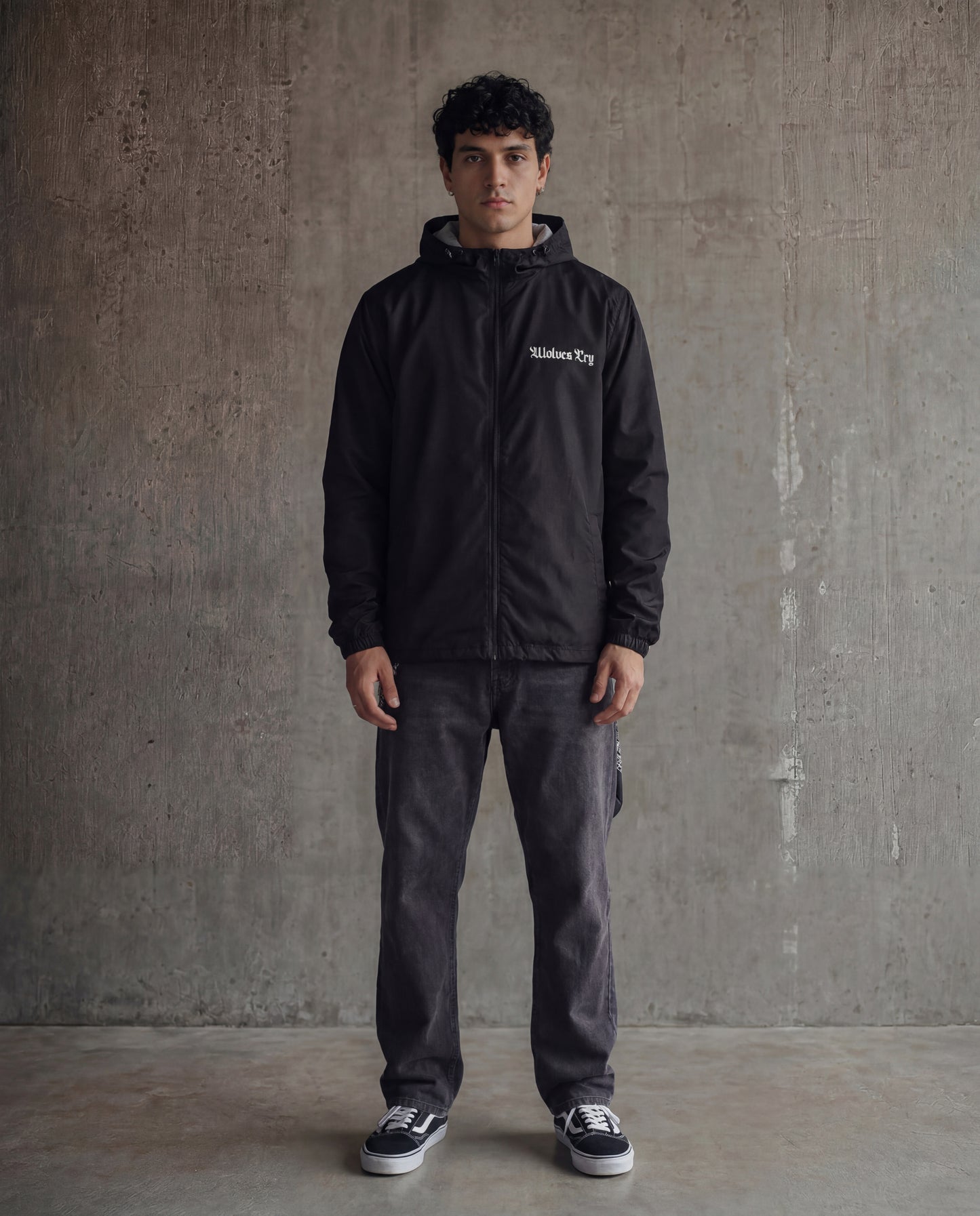 Man wearing a black jacket with a brand logo against a gray concrete wall.