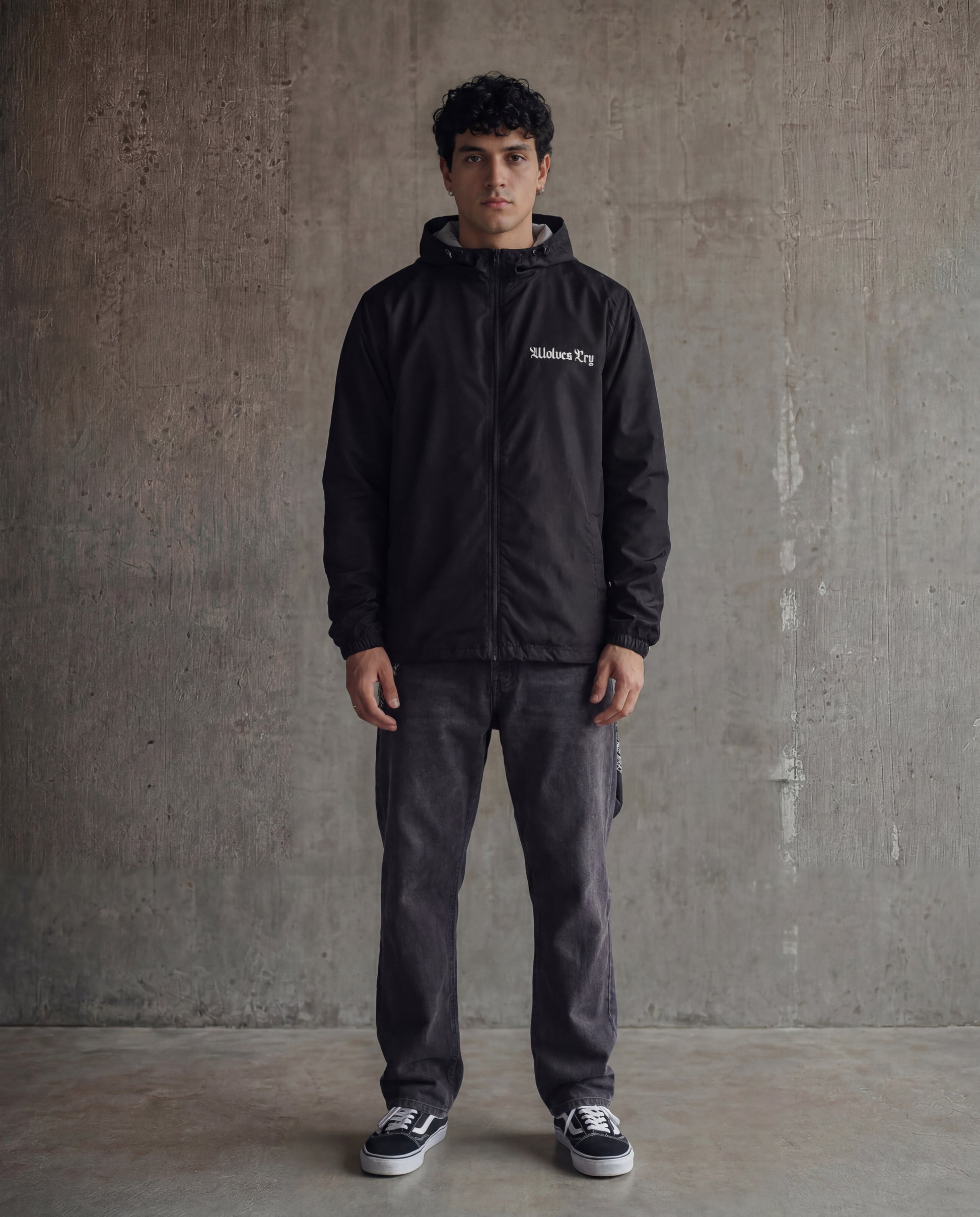Man wearing a black jacket with a brand logo against a gray concrete wall.