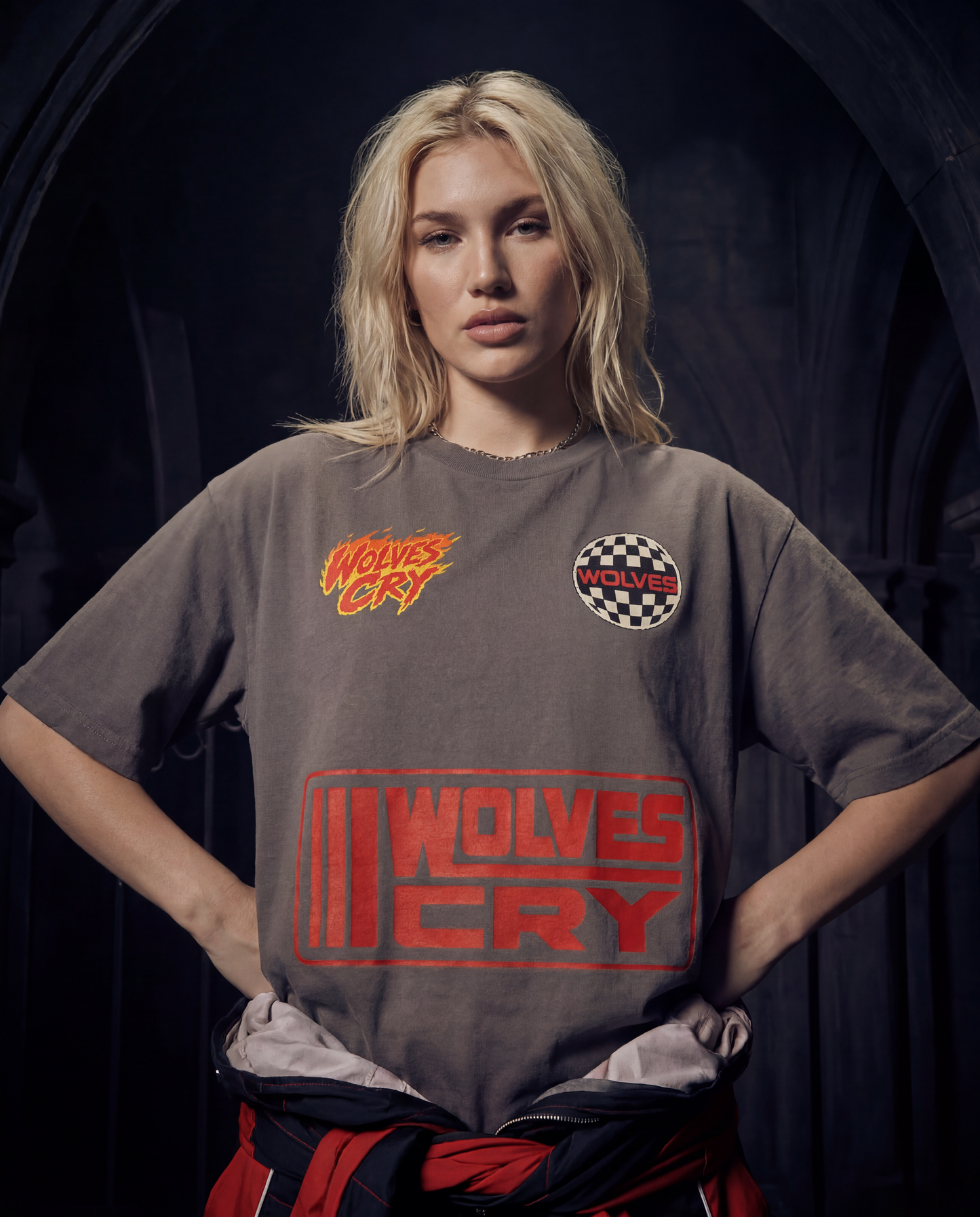 A blonde woman stands with hands on hips wearing a faded dark gray graphic t-shirt featuring the red and orange 'Wolves Cry' logo on the front, set against a dark, moody background.