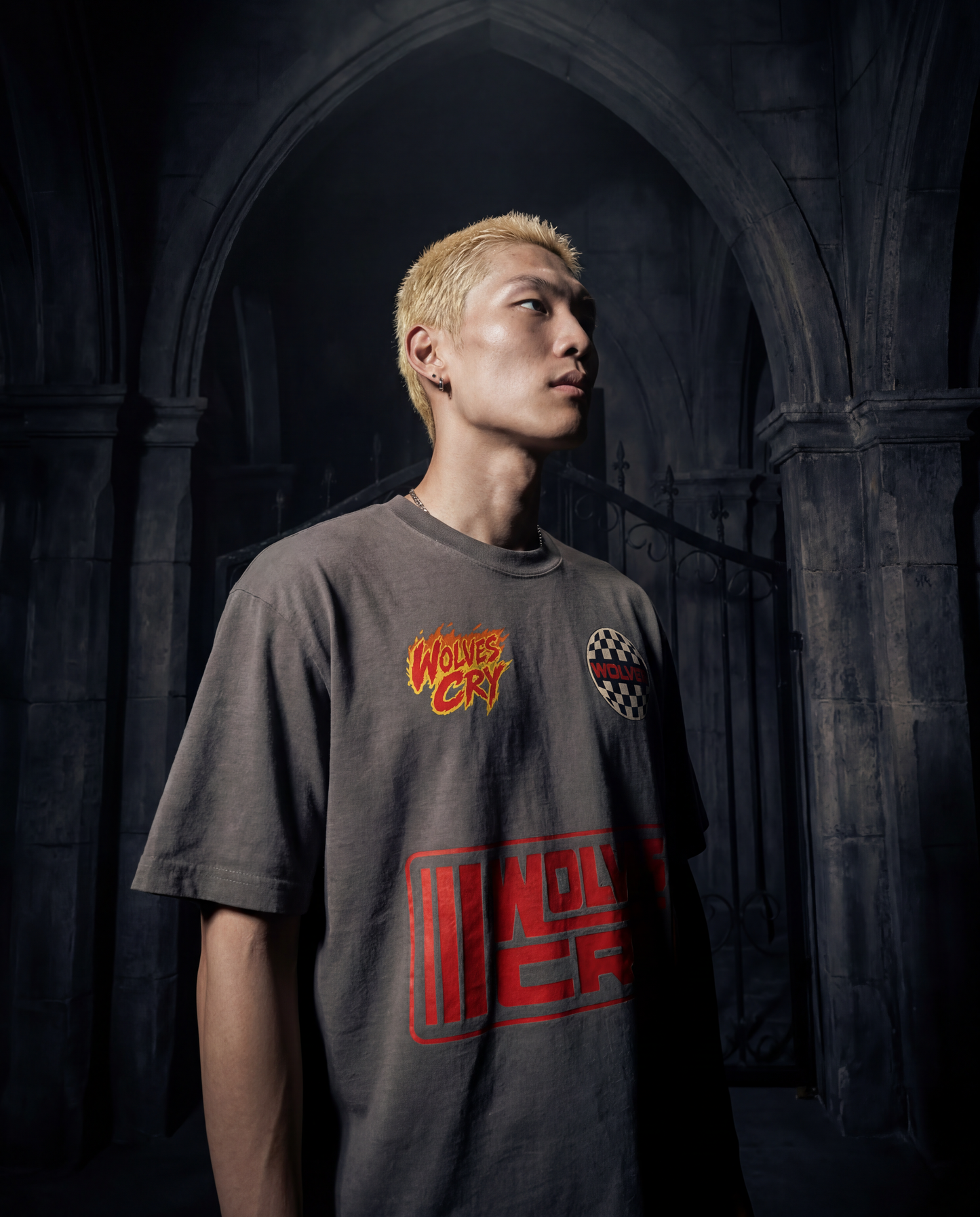 A man with short blonde hair wearing a dark gray wolves cry graphic t-shirt with red and yellow designs, standing in a dark gothic stone archway.