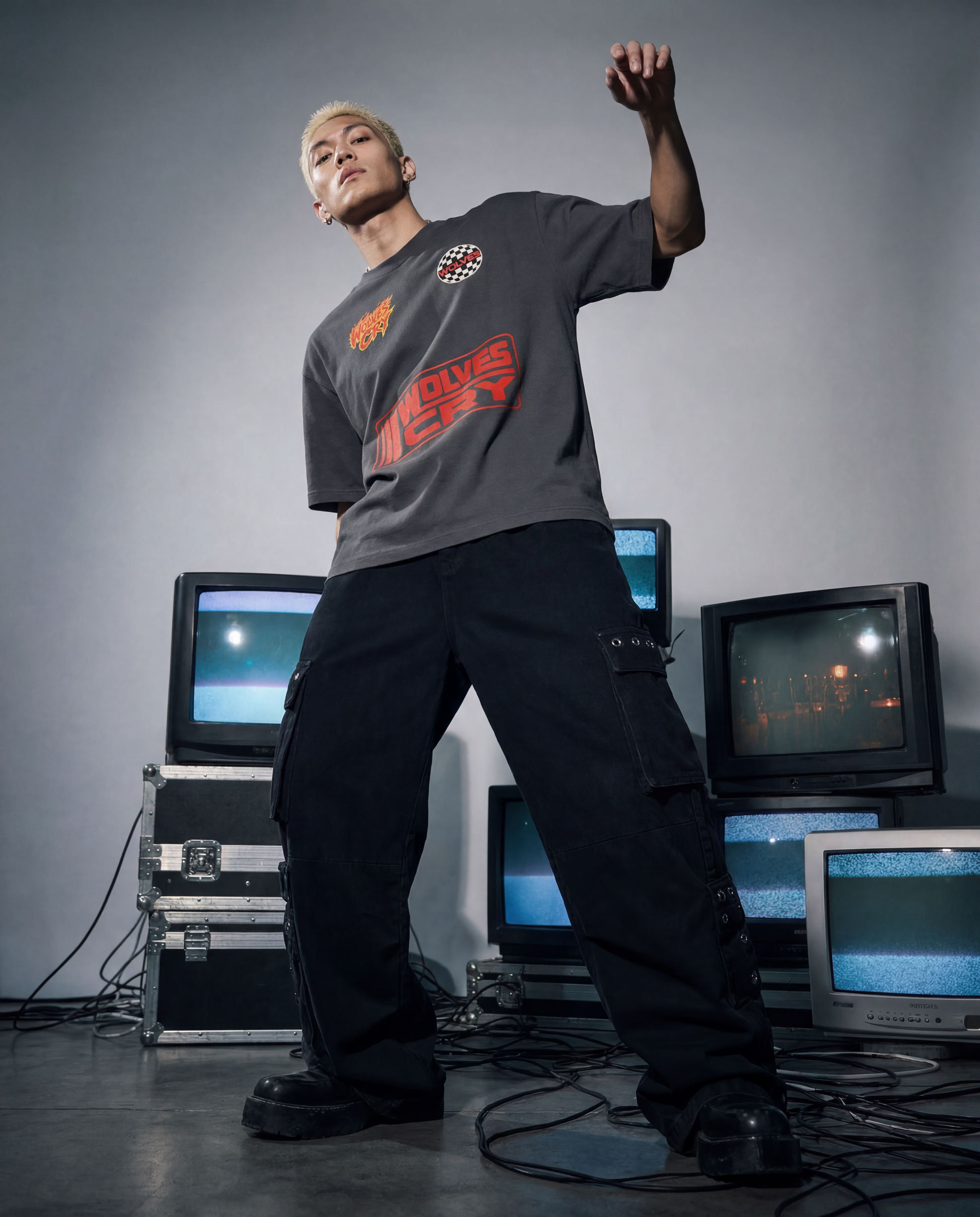 A man wearing a dark gray graphic t-shirt and black cargo pants stands in a studio surrounded by vintage CRT television sets. The t-shirt features orange and red graphics, including the text 'Wolves Cry'.