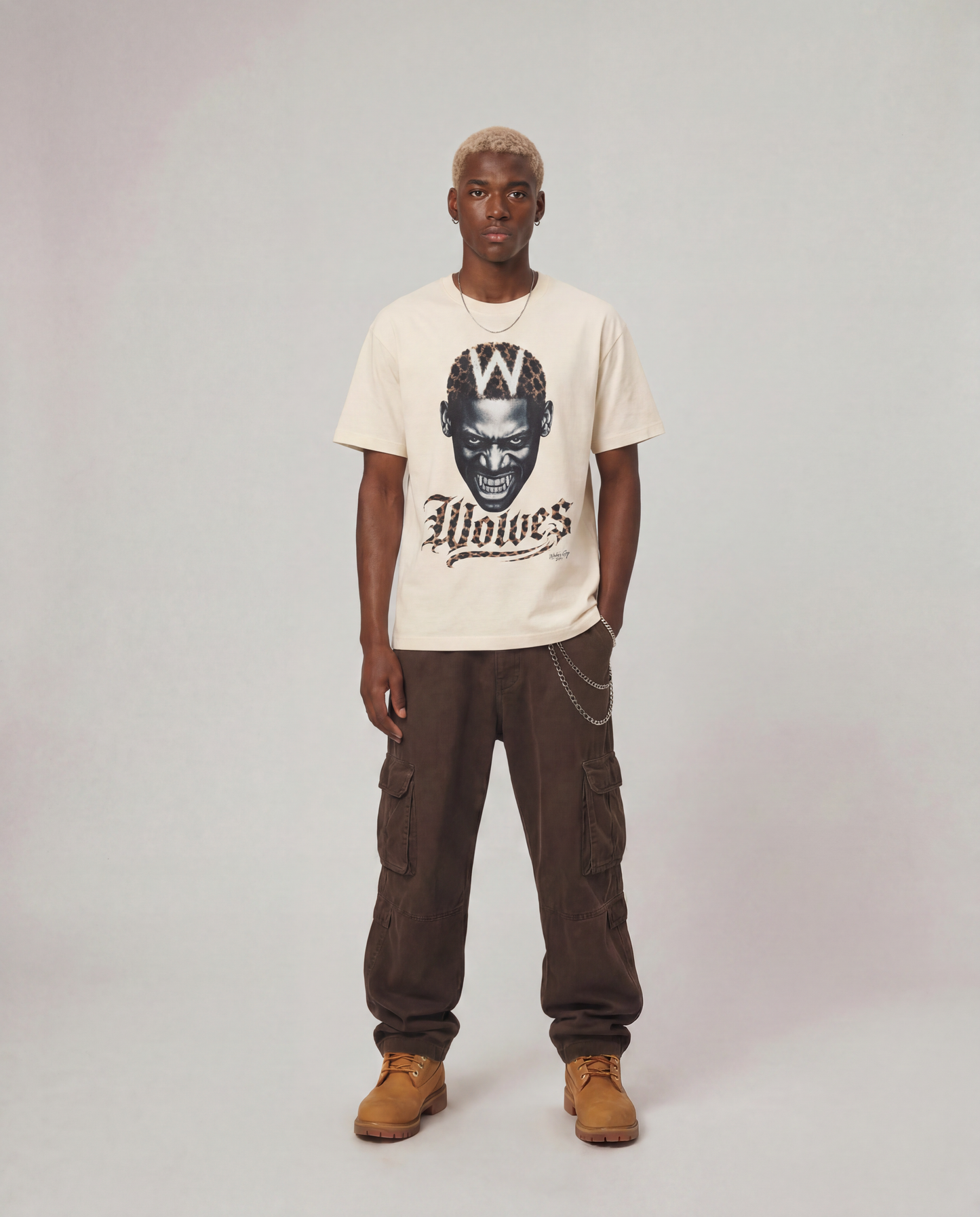 A model stands against a light gray studio background wearing a cream-colored graphic t-shirt with a black print of a wolf head and the word 'Wolves', paired with dark brown cargo pants and tan boots.