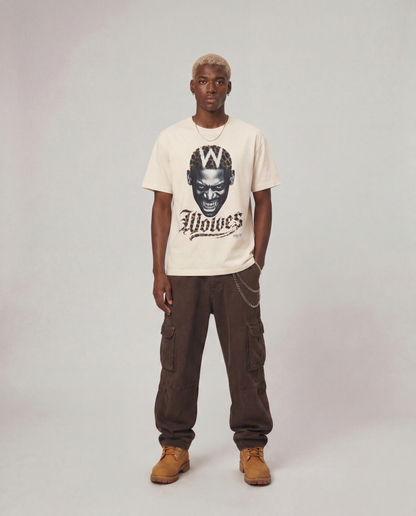 A model stands against a light gray studio background wearing a cream-colored graphic t-shirt with a black print of a wolf head and the word 'Wolves', paired with dark brown cargo pants and tan boots.