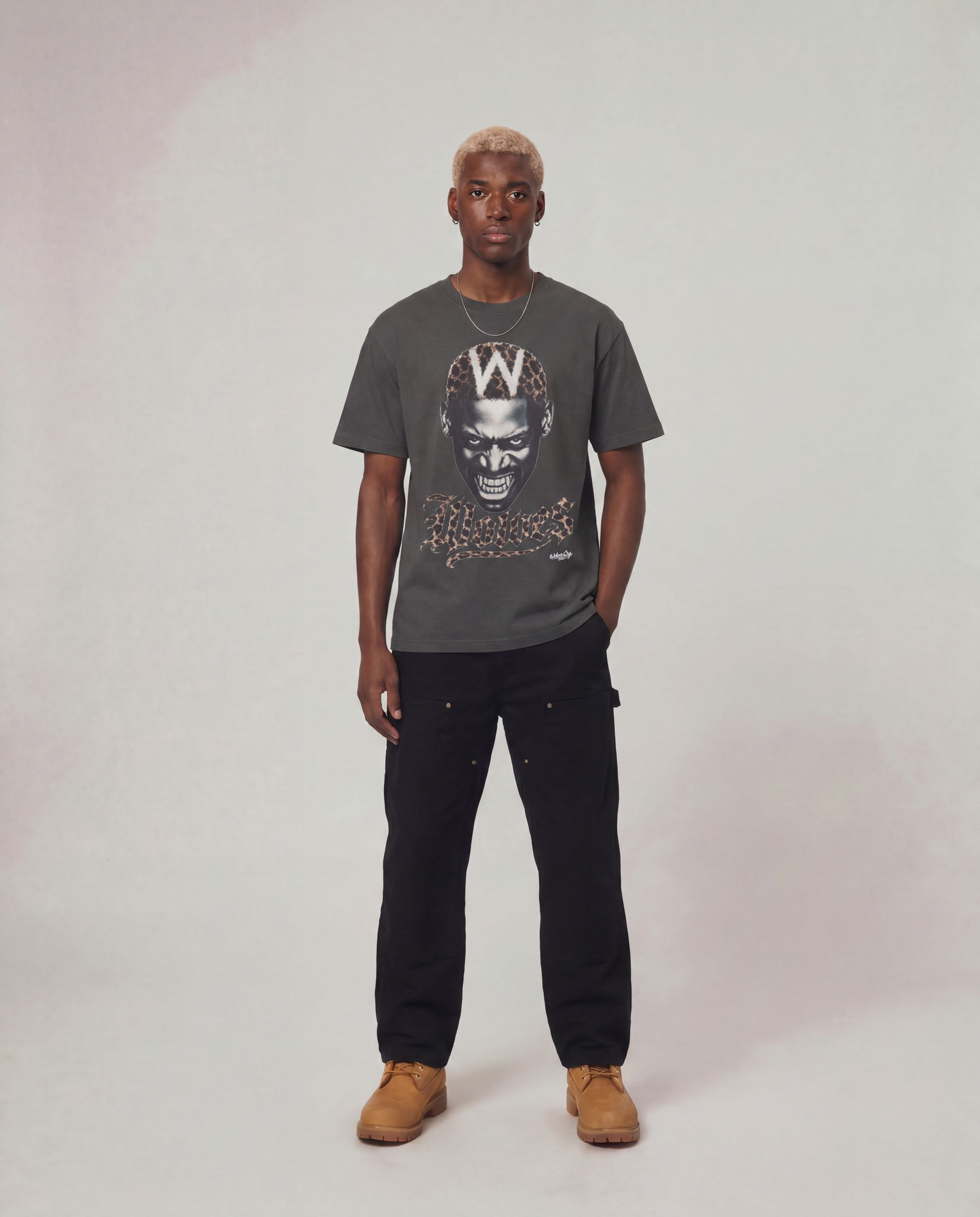 A man modeling a dark gray graphic t-shirt with a stylized face print and a large 'W' on the chest, paired with black cargo pants and tan suede boots, against a light gray studio background.