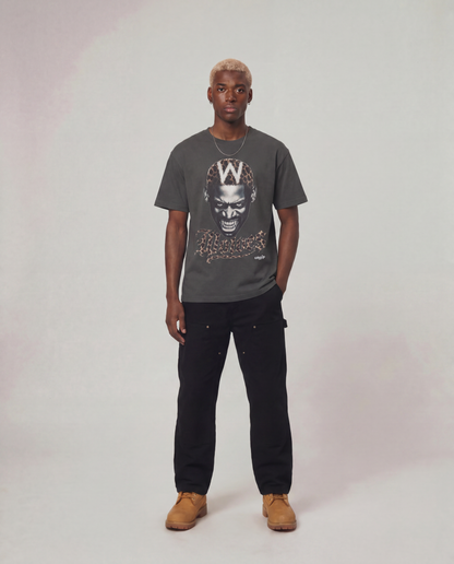 A man modeling a dark gray graphic t-shirt with a stylized face print and a large 'W' on the chest, paired with black cargo pants and tan suede boots, against a light gray studio background.