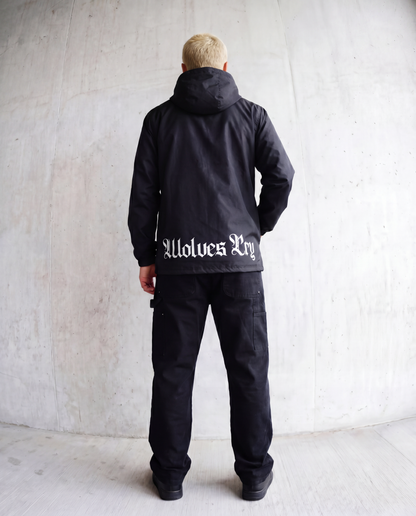 A man viewed from behind wearing a black hooded jacket with 'wolves Cry' printed in white Gothic lettering across the back, paired with black pants, standing against a textured concrete wall.