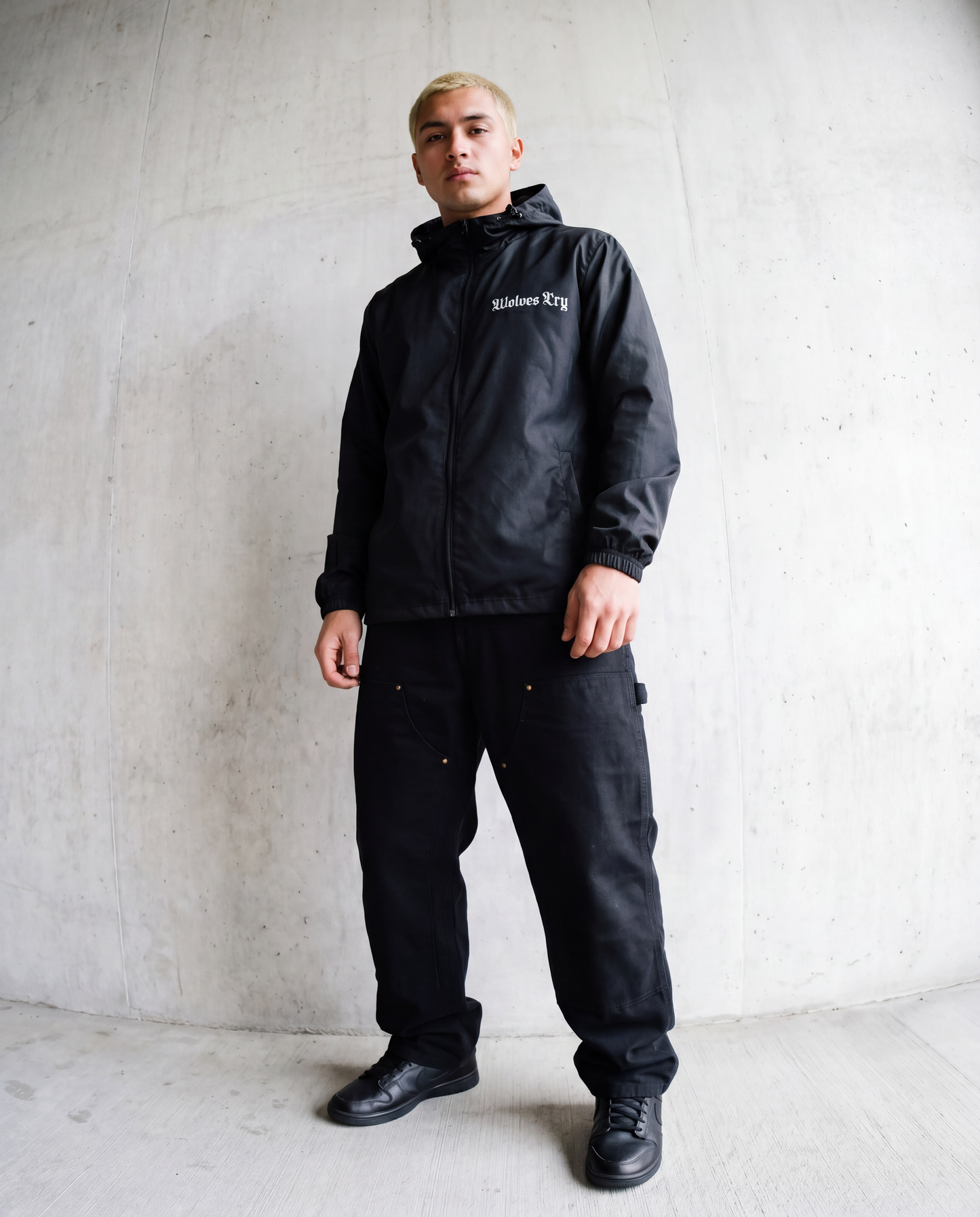 A man wearing a black Wolves Cry hooded jacket and black cargo pants, standing against a textured concrete wall.