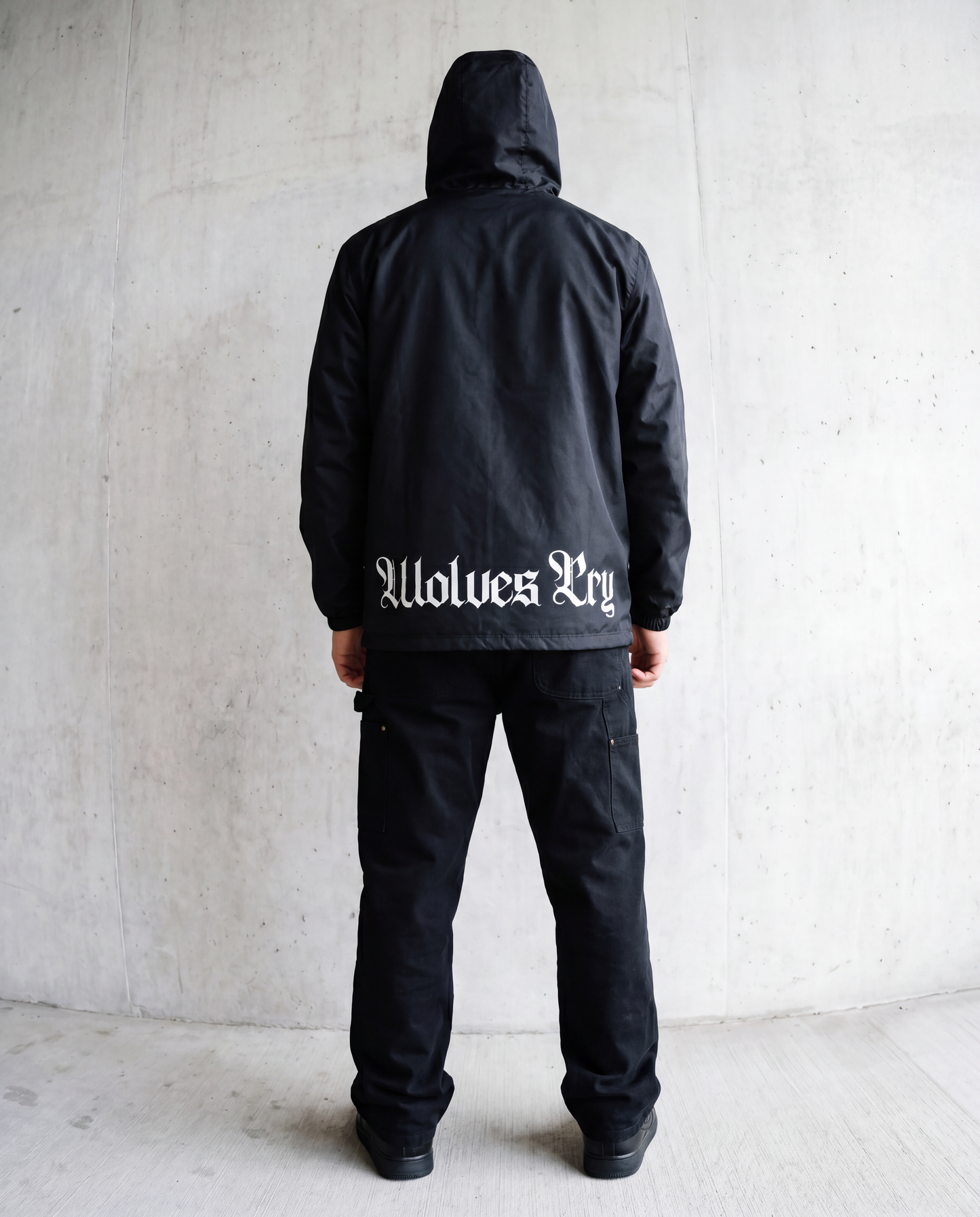 A person standing with their back to the camera, wearing a black hooded jacket with 'Wolves Cry' printed in white Gothic lettering across the lower back, paired with black pants, set against a concrete wall background.